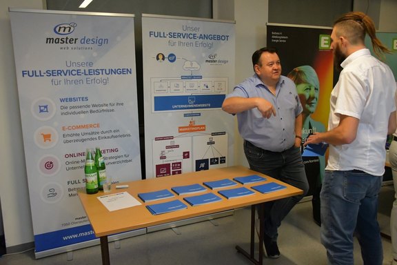 Mitarbeiter von master design im Gespräch mit einem Besucher beim Karrieretag 2025 an der HTL Pinkafeld, Stand mit Informationsmaterial.